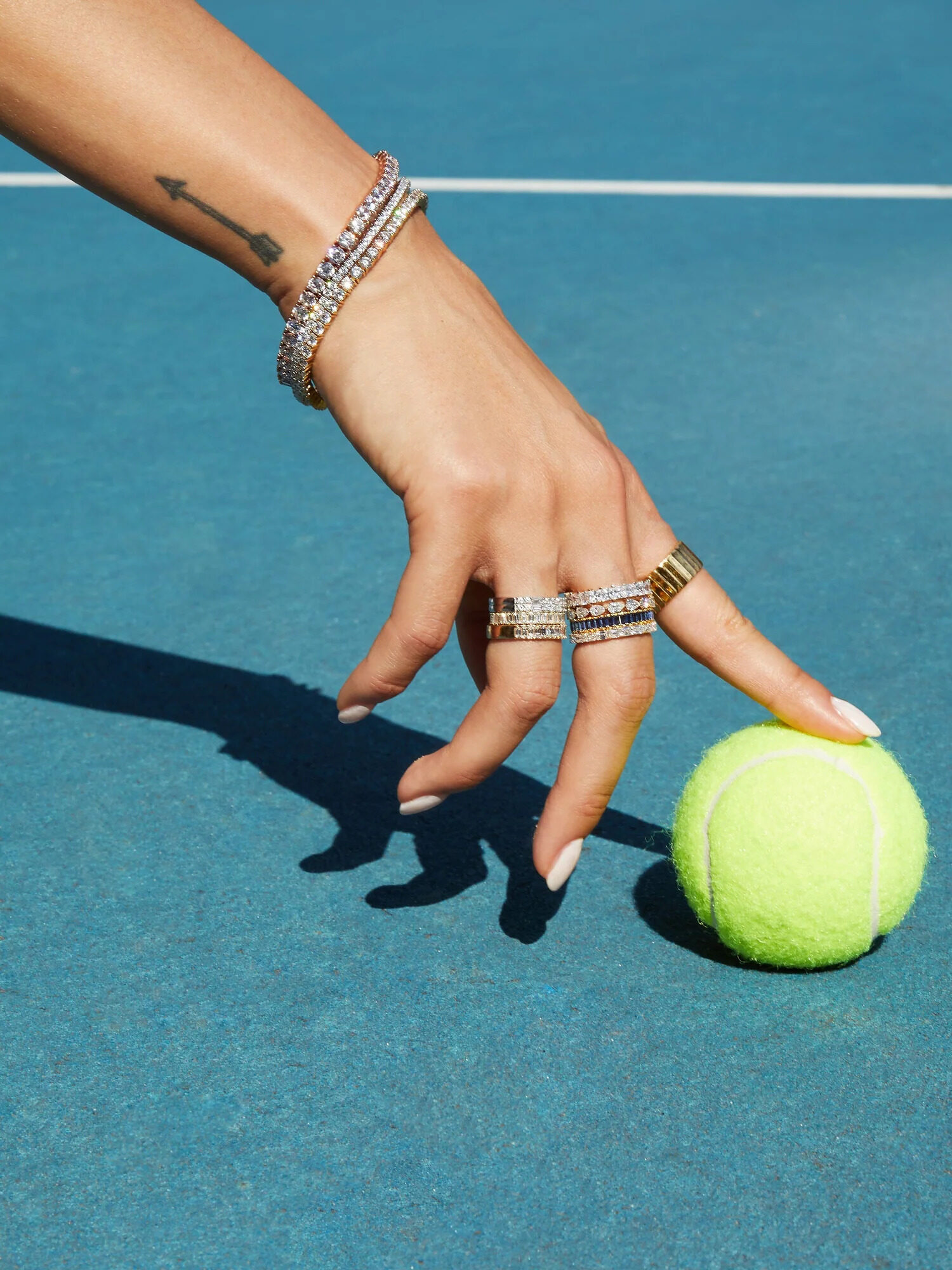 A close up shot of a hand on a tennis court touching a tennis ball with their index finger while wearing a variety of silver and gold Aurate rings and tennis bracelets.
