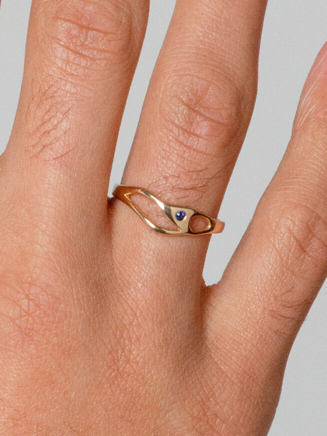 A hand shot of a curvet cut out gold ring with a tiny blue stone embedded by Bario Neal.