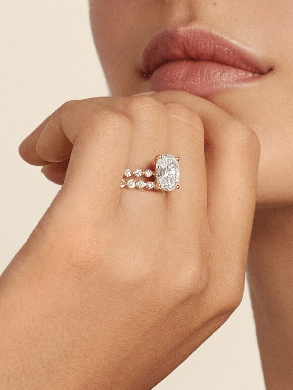 A close up studio shot of a model wearing a diamond oval solitaire engagement ring with a scalloped diamond band by Brilliant Earth.
